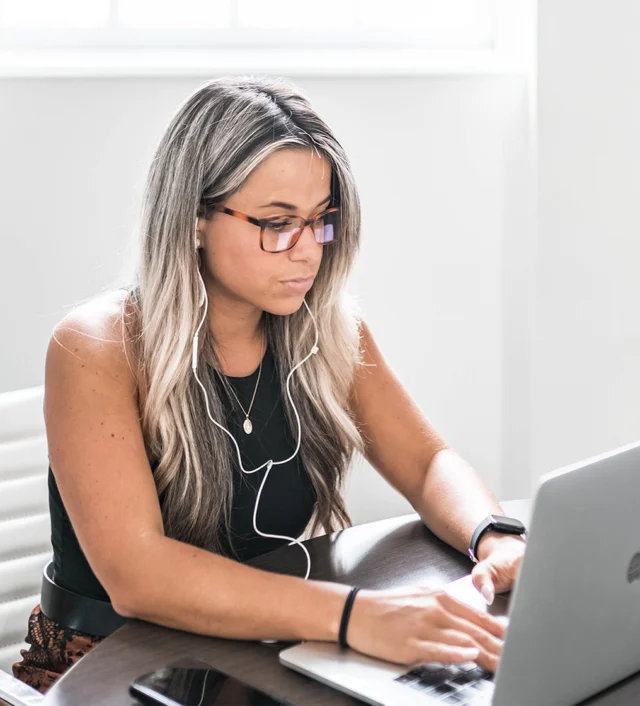 Woman with long hair and glasses working on a laptop at a desk, wearing earphones.