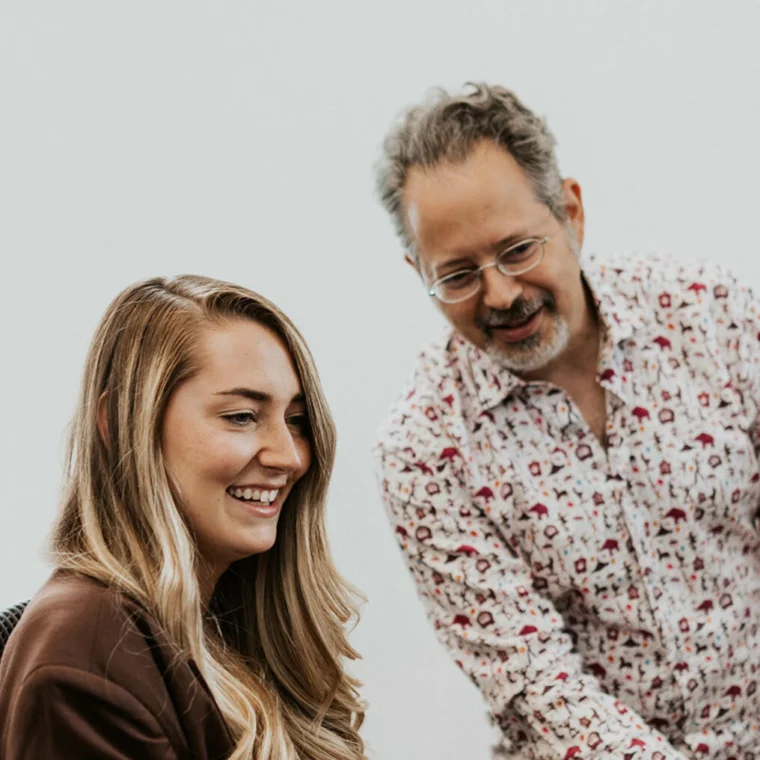 Two people are engaged in conversation. The woman is seated and smiling, while the man stands beside her, leaning slightly forward.