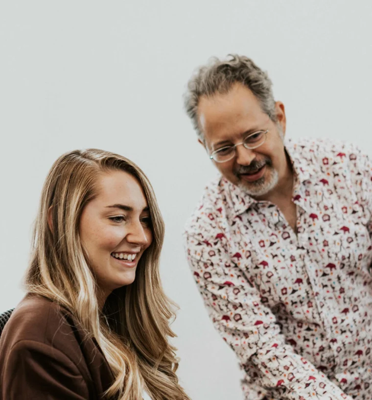 Two people are engaged in conversation. The woman is seated and smiling, while the man stands beside her, leaning slightly forward.