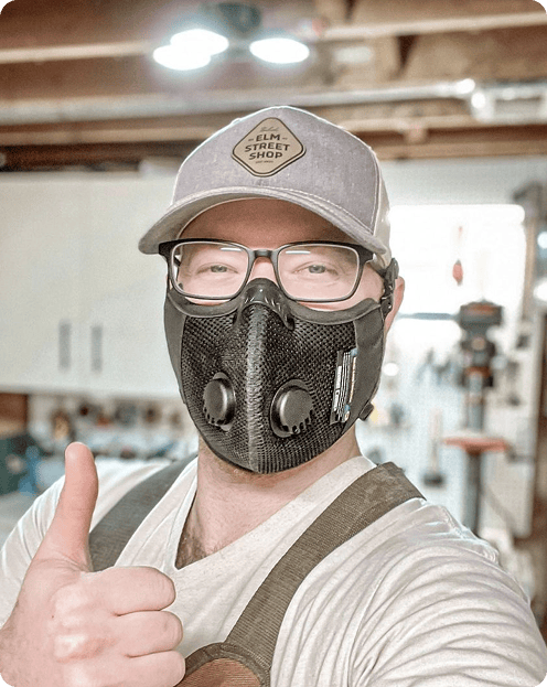 Person wearing a cap, glasses, and a ventilation mask gives a thumbs-up in a workshop setting.