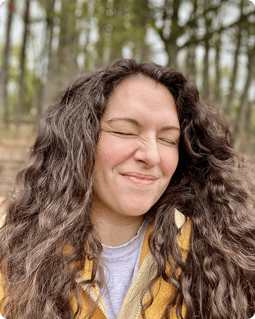 Person with long curly hair smiles with eyes closed in a wooded area. They are wearing a yellow jacket and white shirt.