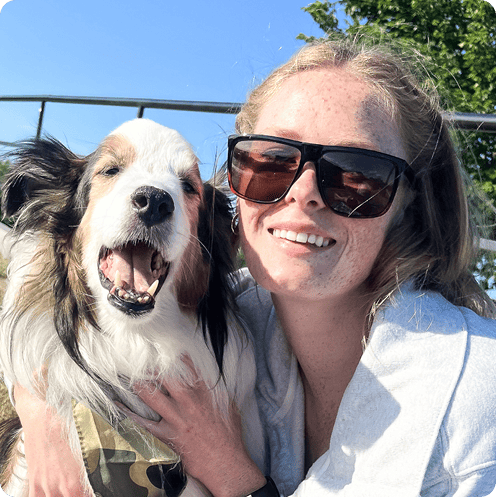 A woman wearing sunglasses smiles while holding a yawning dog in her arms outside on a sunny day.