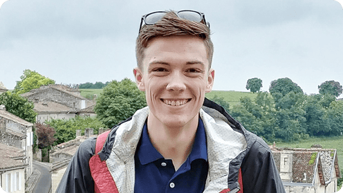 Young person smiling, standing outdoors with trees and buildings in the background, wearing a jacket and sunglasses on their head.