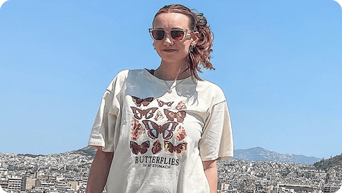 Person wearing a butterfly-patterned T-shirt and sunglasses, standing outdoors with a city and mountains in the background on a clear day.