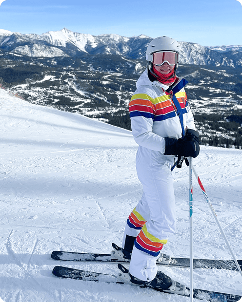 A person in a colorful ski suit, helmet, and goggles stands on skis on a snowy mountain slope with a mountainous landscape in the background.