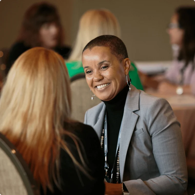 A smiling person in a gray blazer sits at a table, engaged in conversation with another person with long hair. Other individuals are visible in the background.