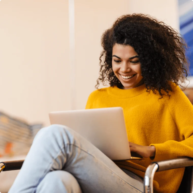 Person with curly hair in a yellow sweater is smiling while using a laptop on their lap, seated on a chair indoors.