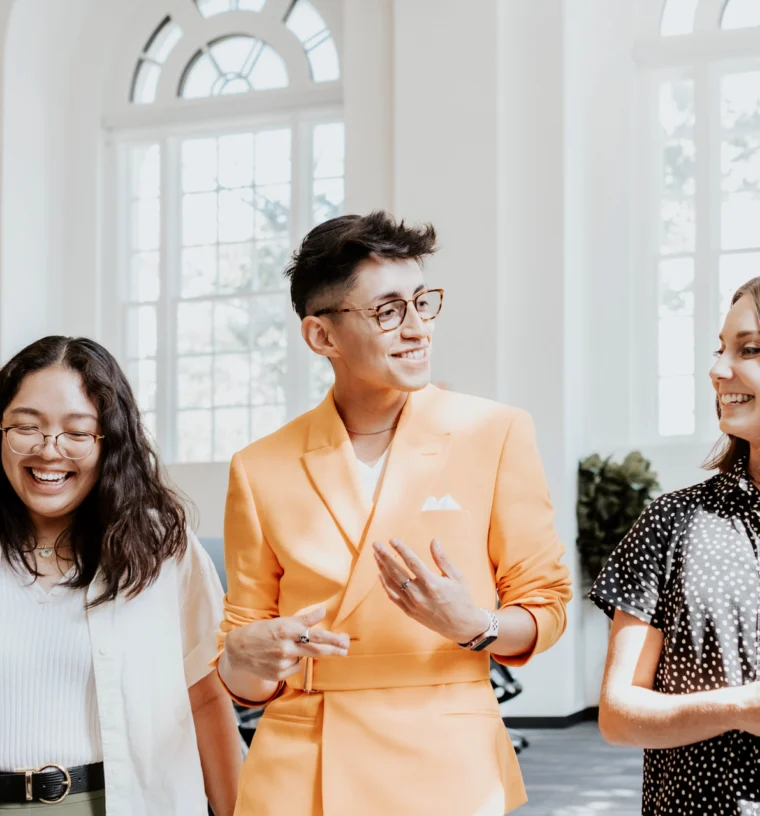 Three people stand indoors, conversing and smiling. One wears an orange suit, while the others wear casual attire. Large windows are visible in the background.