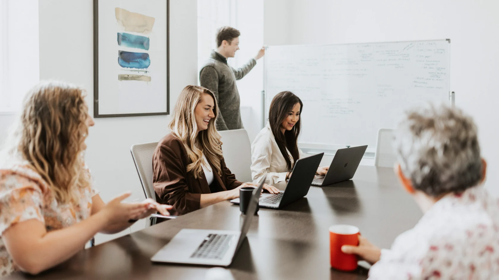 A group of people in a meeting room are discussing around a table. Two people are using laptops, and one person is writing on a whiteboard.