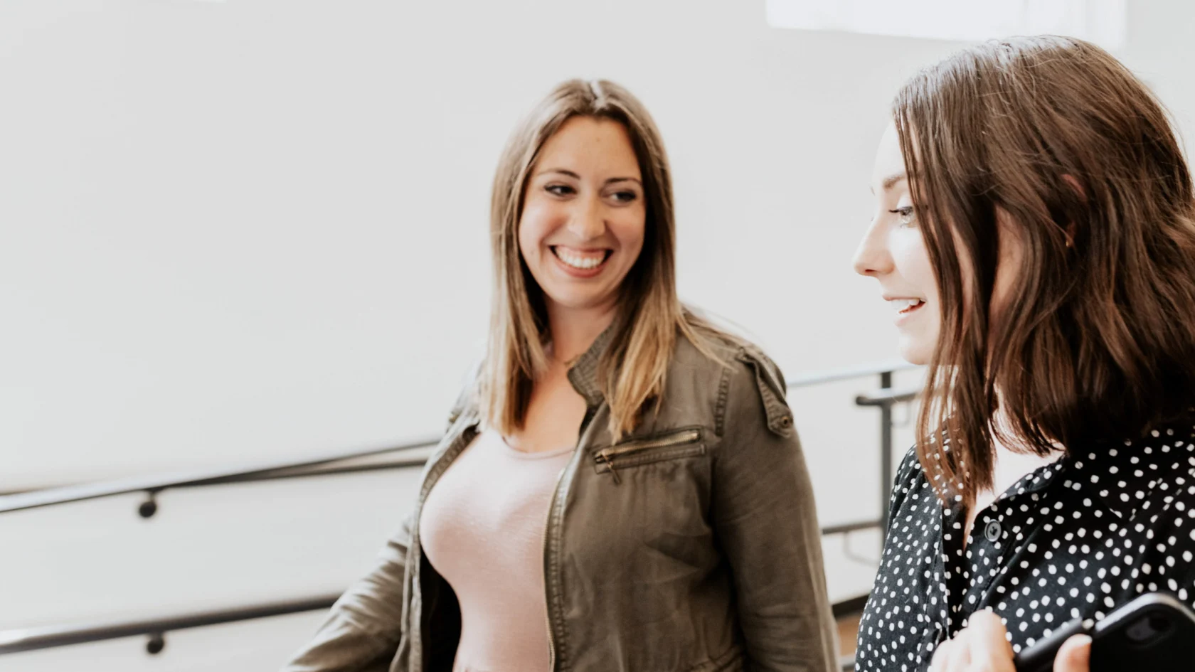 Two women walking indoors, one smiling at the other. Both dressed casually, one in a jacket, the other in a polka dot top. Background shows light-colored walls and railings.