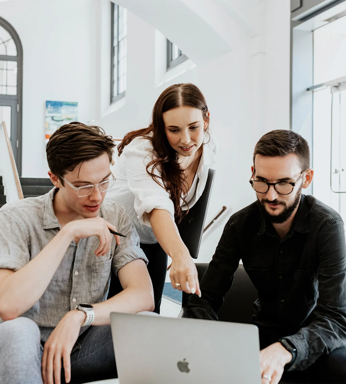 Three people collaborate, looking at a laptop screen in a bright office space.