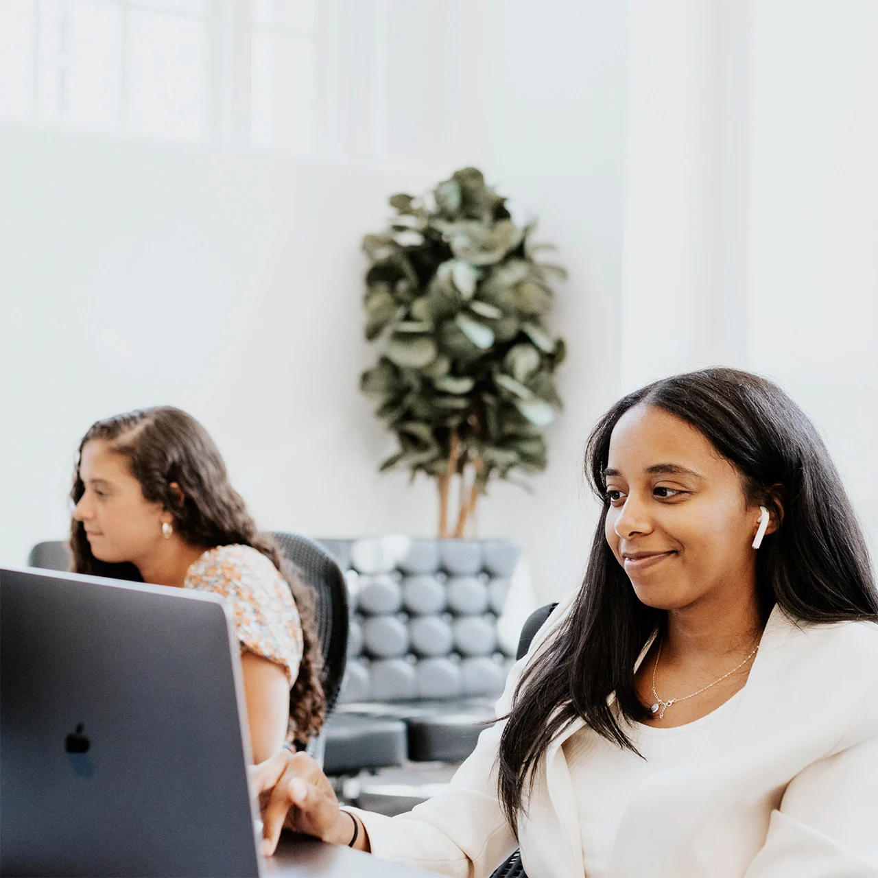 Two people work on laptops in an office. One is in focus, smiling and wearing wireless earphones. A plant and couch are in the background.