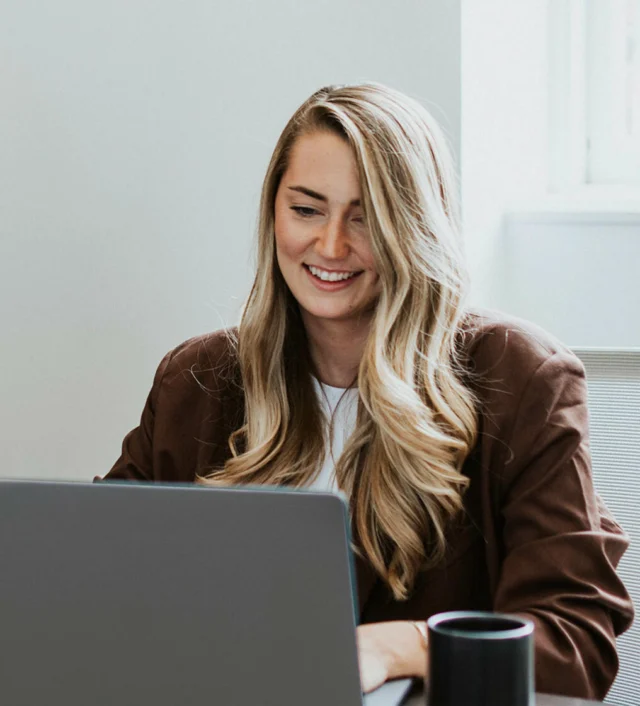 A person with long hair sits at a desk, smiling while looking at a laptop. A mug is next to the laptop.