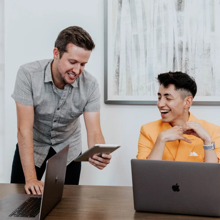Two people at a desk with laptops; one standing, showing a tablet, and the other seated, smiling.