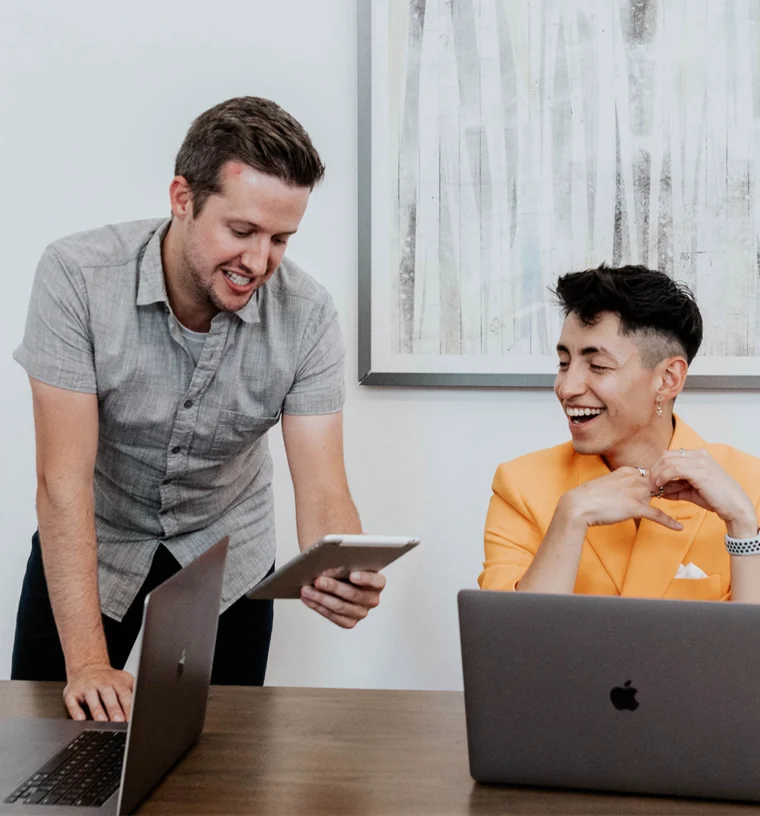 Two people at a desk with laptops; one standing, showing a tablet, and the other seated, smiling.