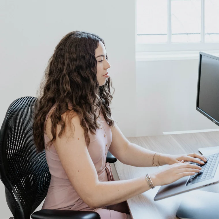 Person with long curly hair typing on a computer keyboard, seated at a wooden desk in a bright room with a window.