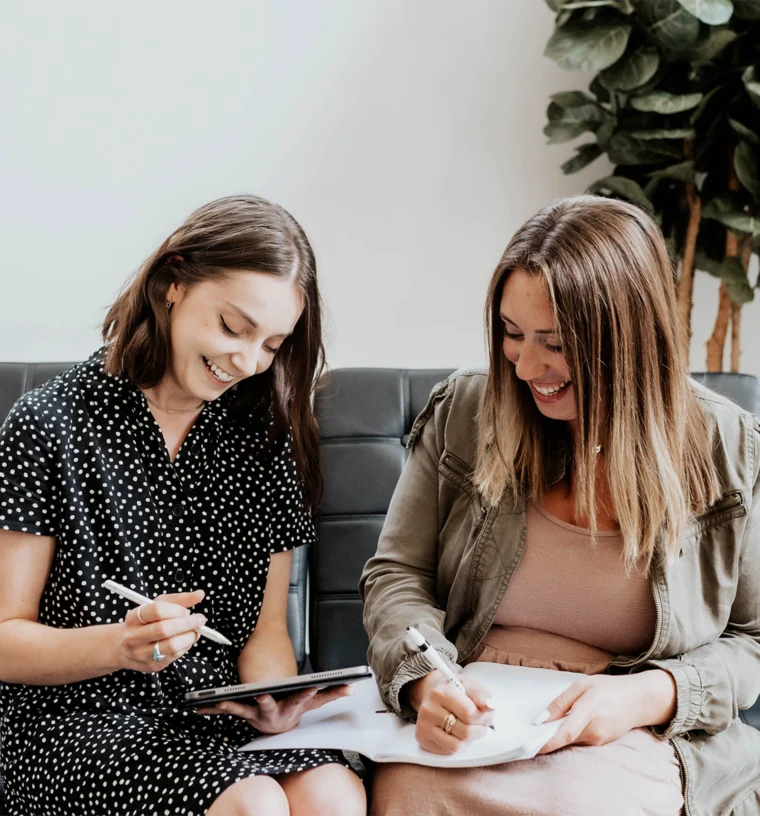 Two people are sitting on a sofa; one is writing in a notebook, and the other is using a tablet. They are smiling and appear to be having a friendly discussion.
