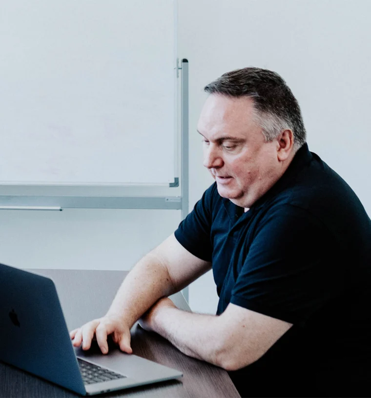 A person in a black shirt sits at a table, looking at a laptop, in front of a whiteboard.