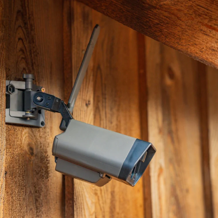 A security camera with an antenna is mounted on a wooden wall under a slanted roof.