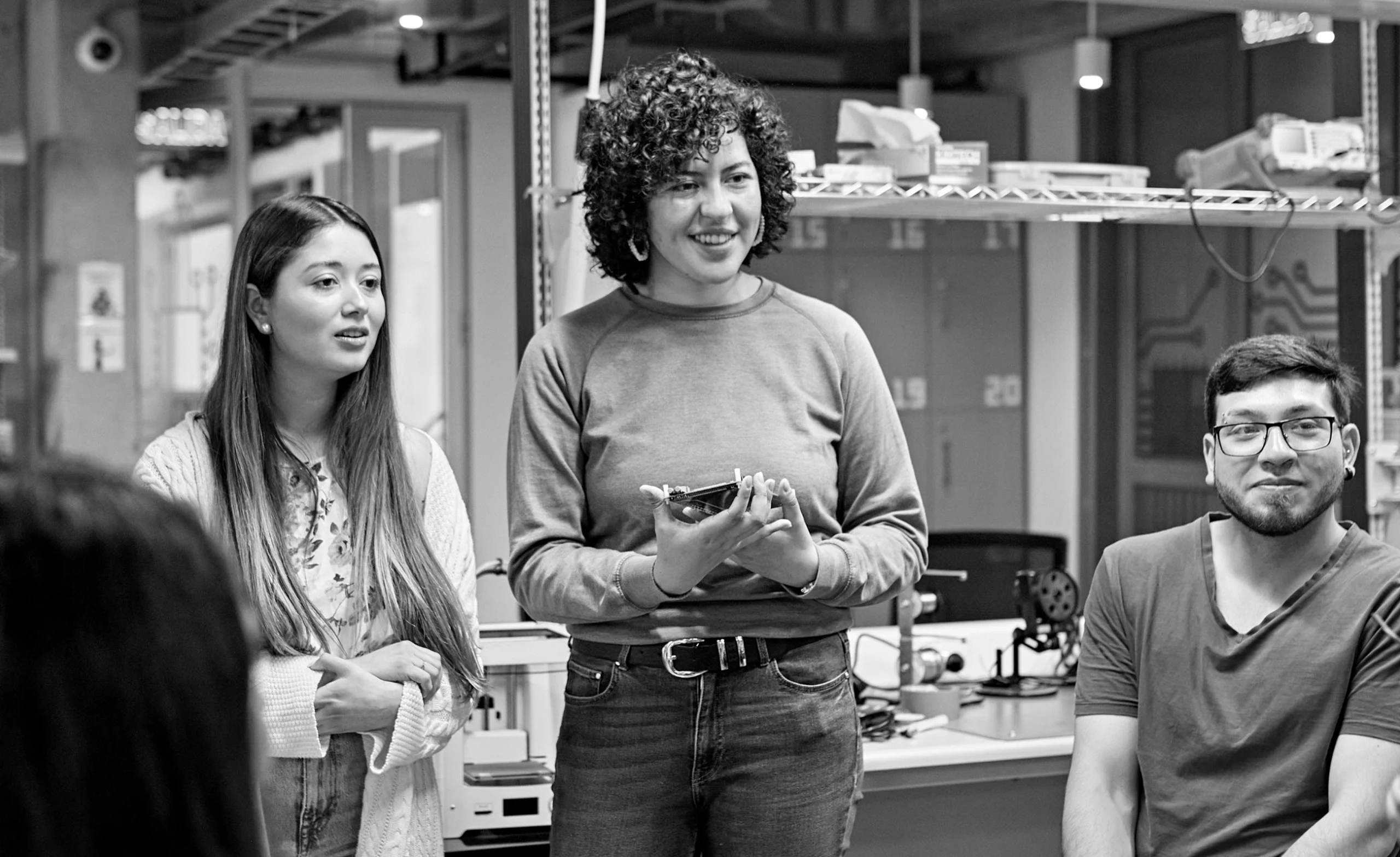 Three people in a workspace, one speaking while holding an item, others listening. Shelves and equipment are visible in the background. Black and white image.