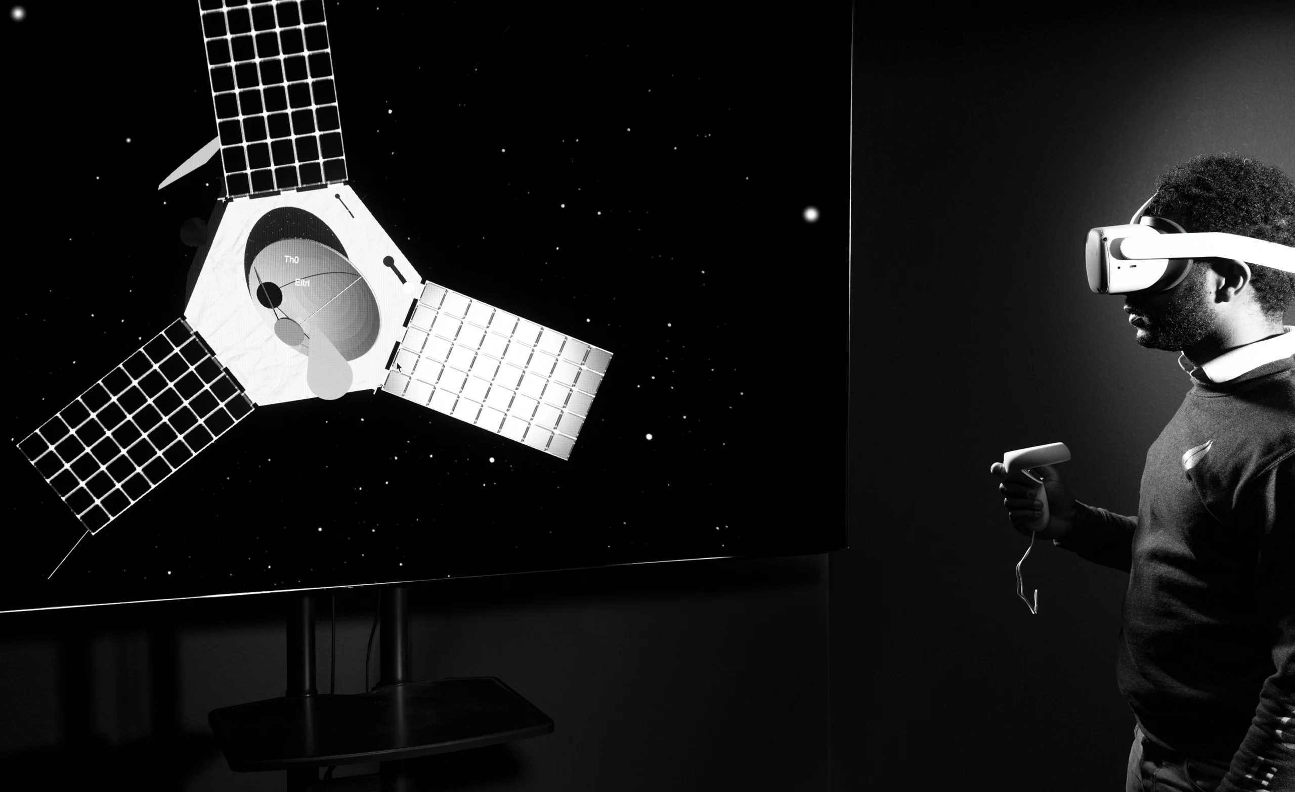 A man wearing VR goggles holds a controller, standing in front of a screen displaying a satellite in space.