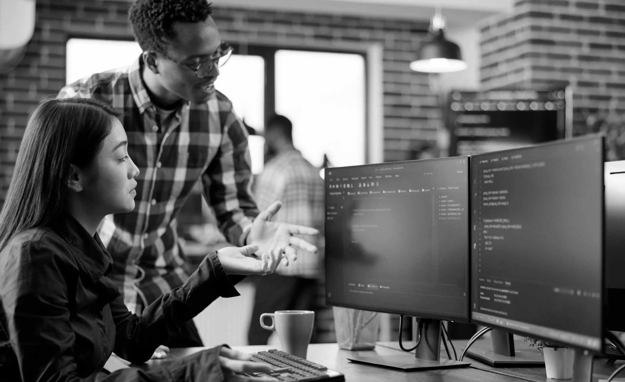 Two people in an office setting are engaged in a discussion in front of dual computer monitors displaying code. The workspace has a modern design with large windows and brick walls.