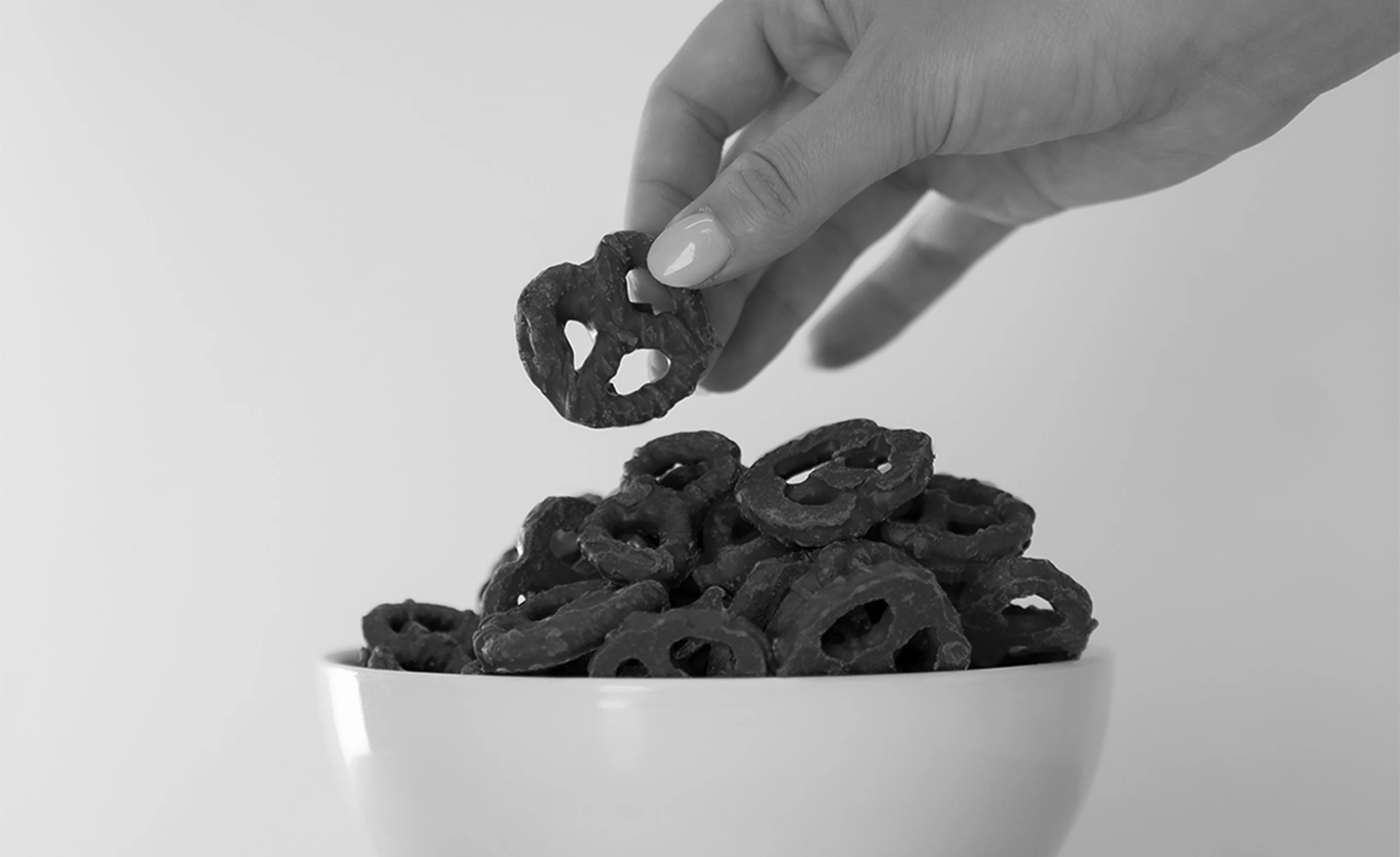 A hand reaches into a bowl of chocolate-covered pretzels, holding one above the bowl. The image is in black and white.
