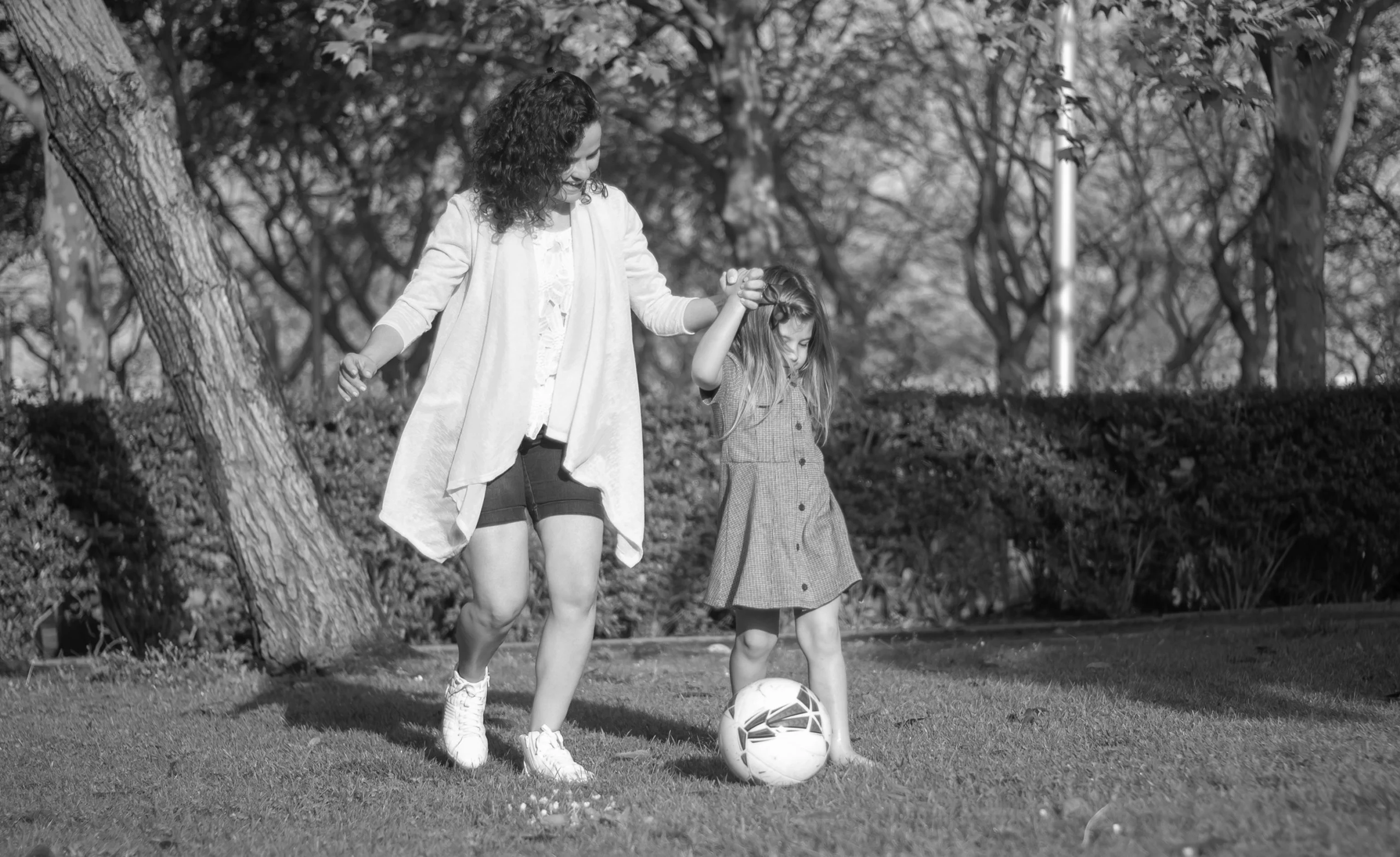 Woman and child playing with a soccer ball on a grassy area, surrounded by trees.