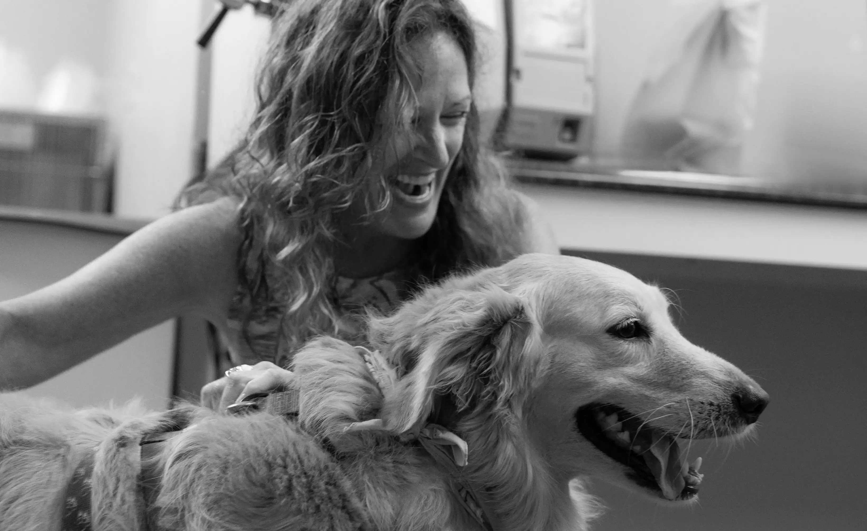 A woman with curly hair smiles and pets a golden retriever indoors. The image is in black and white.