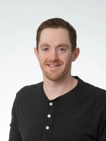 A man with short red hair and a trimmed beard wears a black henley shirt and smiles at the camera against a plain white background.